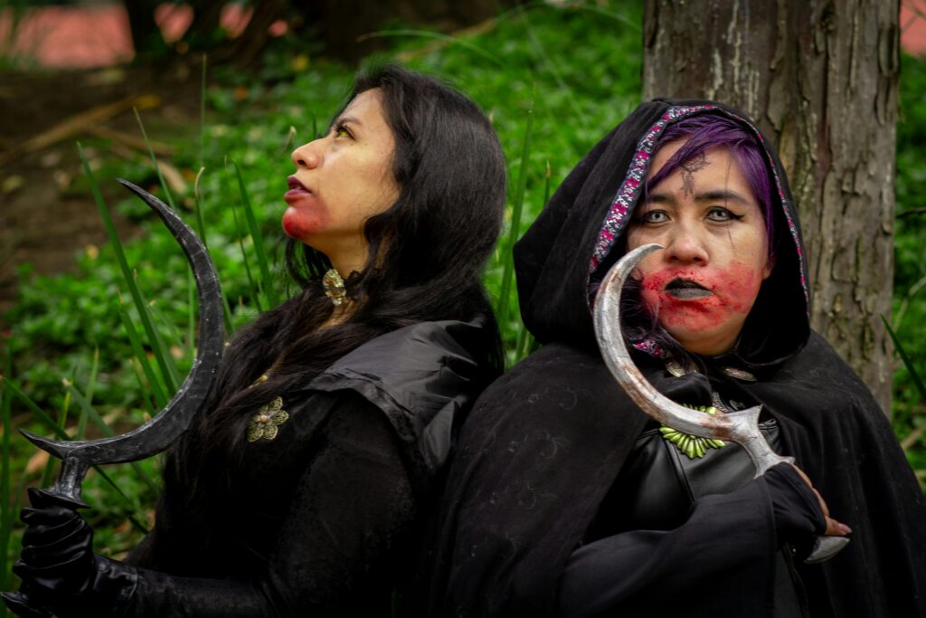 Two adults in dark, dramatic cosplay with makeup and props in a forest setting.