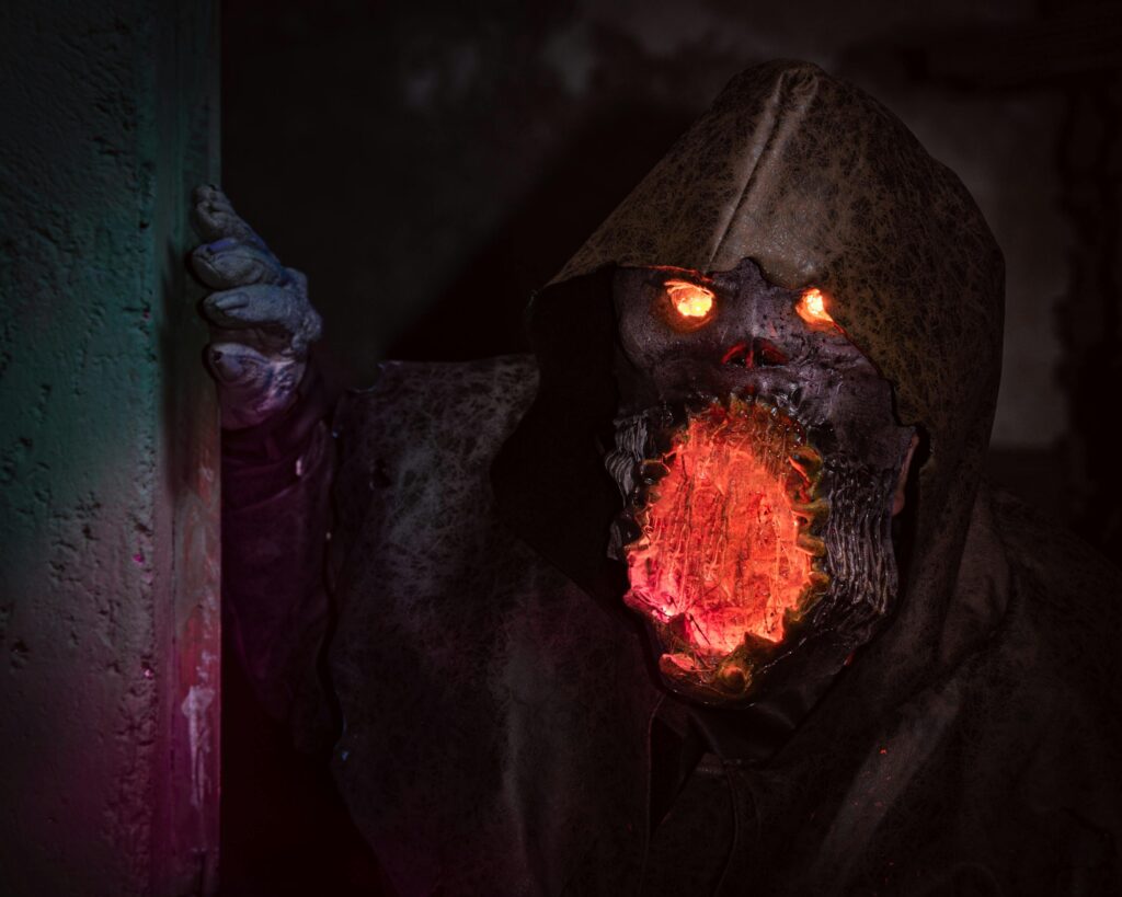 A menacing monster with glowing eyes and mouth in detailed horror cosplay peeking from a shadowy corner.