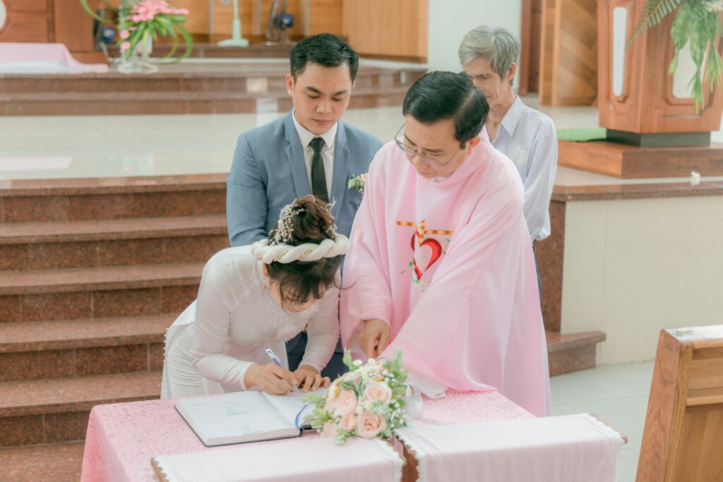 Bride and groom signing marriage documents with priest during church ceremony.