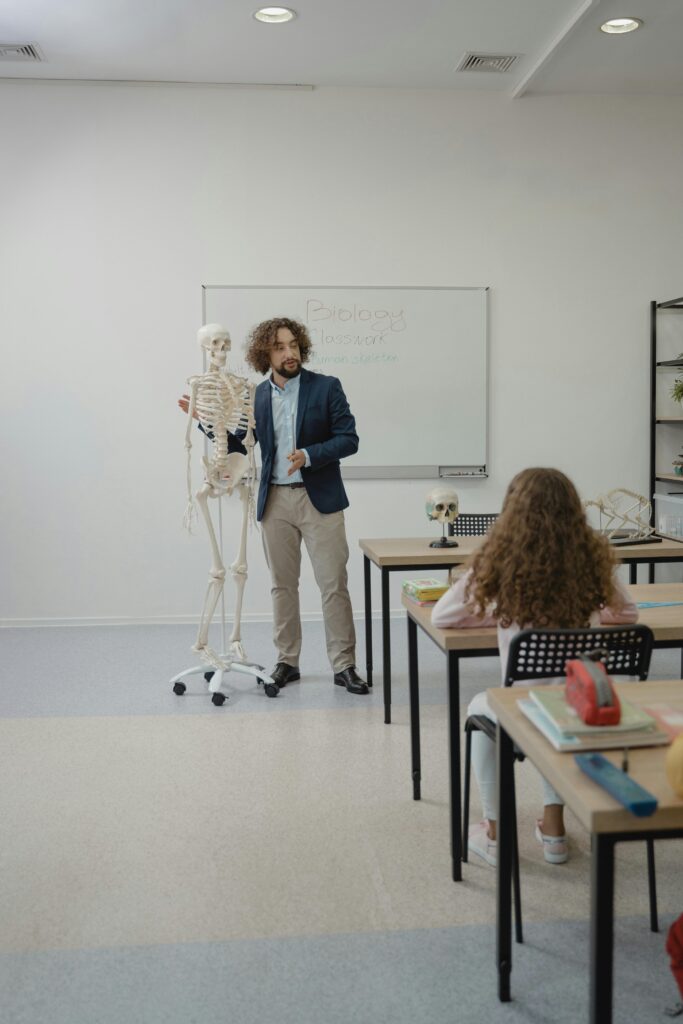 Education System United States and India Teacher explaining anatomy with skeleton model in bright classroom setting.