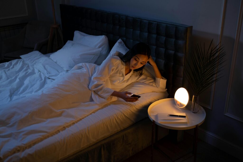 Insomnia in the United States An Asian woman on a bed using her phone in a dim bedroom, reflecting on insomnia or night routines.