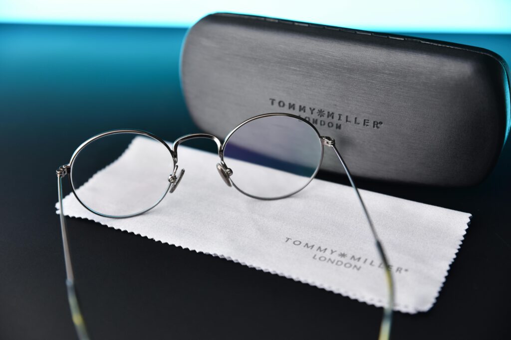 Close-up of stylish Tommy Miller eyeglasses with branded case and cleaning cloth on a dark surface.