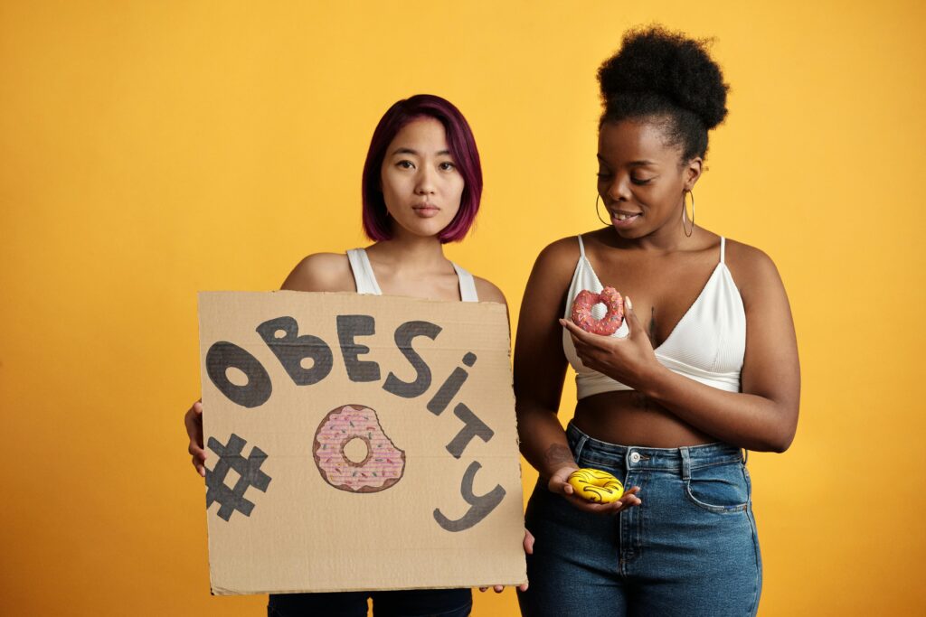 Two women discussing obesity awareness, holding donuts and a sign, against a yellow background.