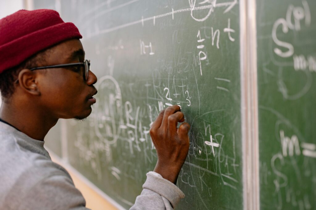A young man with eyeglasses and a red beanie solving math problems on a chalkboard.